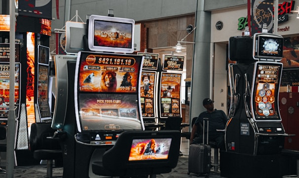 A collection of slot machines is situated inside a building with bright, colorful screens displaying various games and gaming themes. A person is seated nearby, seemingly relaxed with luggage beside them, and the atmosphere is lively with electronic displays.